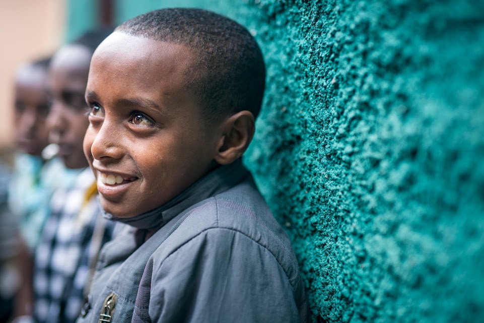 Boy in Ethiopia sits against a blue wall smiling upward