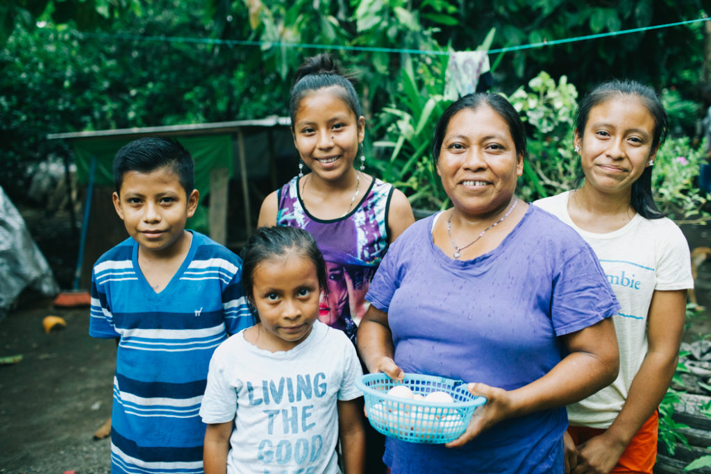 A mother in Guatemala holds a basket of eggs while smiling with her family
