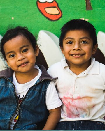 A boy and girl in Guatemala smile, sitting against a green wall