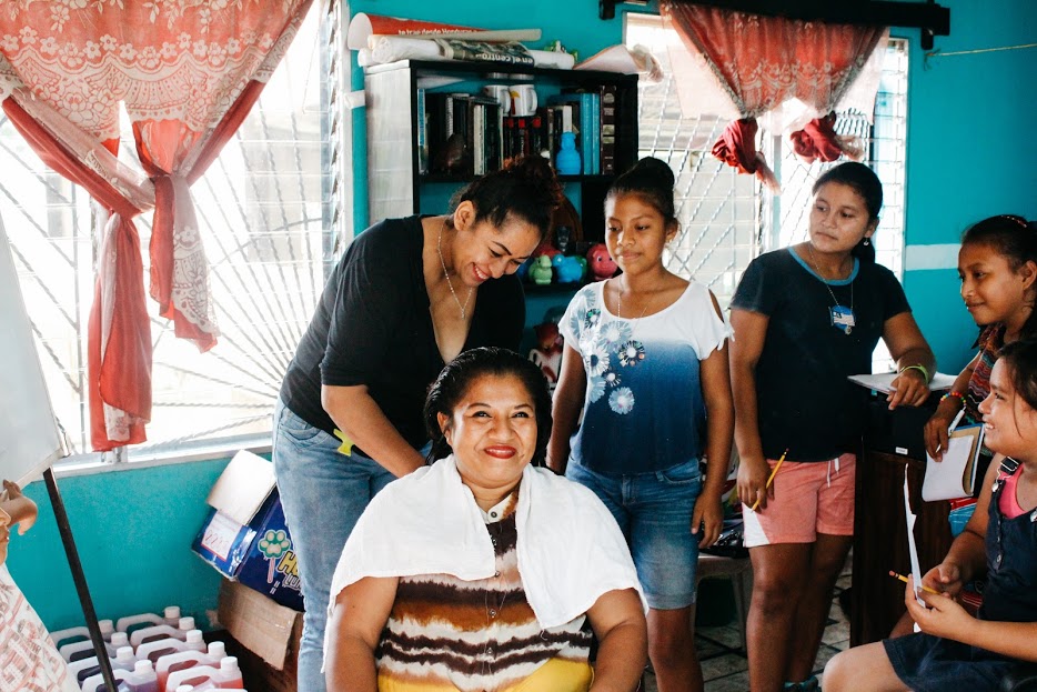 A woman sits in the middle of a circle of girls, getting her hair cut