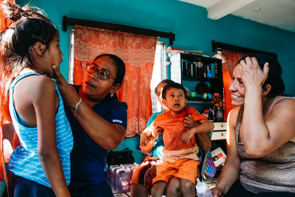 A woman holds a girl's face to perform a medical exam while the girl's family sits nearby