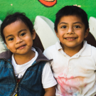 A boy and girl in Guatemala smile, sitting against a green wall